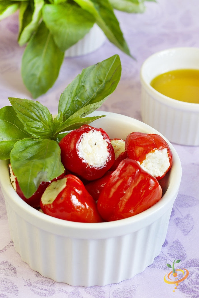 Red cherry hot peppers stuffed with cream cheese in a white ramekin with fresh basil leaves and olive oil in the background